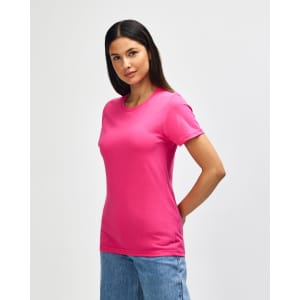 A pink Gildan Softstyle ladies midweight t-shirt displayed on a model, wearing blue jeans, with a plain background.
