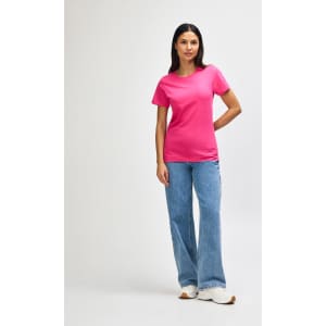 A pink Gildan Softstyle ladies midweight t-shirt, worn with blue jeans and white sneakers. The model stands against a plain background.