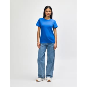 A woman stands wearing a blue Gildan Softstyle ladies midweight T-shirt and jeans, paired with white sneakers.