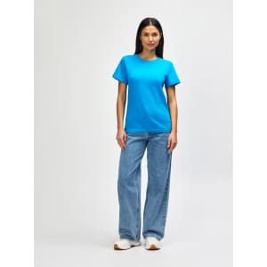 A woman stands wearing a teal Gildan Softstyle Ladies Midweight T-Shirt paired with blue jeans and white sneakers.