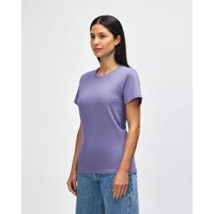 A woman wearing a purple Gildan Softstyle Ladies Midweight T-Shirt paired with blue jeans, smiling at the camera.
