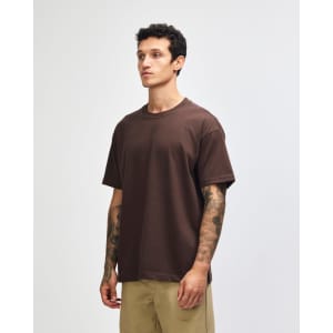 A brown heavyweight t-shirt is held by a model wearing green cargo pants. The shirt has a logo on it.