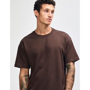 A brown super heavyweight t-shirt on a model with tattoos, featuring a plain design and a logo on the garment.