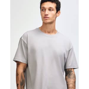 A grey American Apparel Super Heavyweight T-Shirt displayed on a model with curly hair. The shirt has a logo.