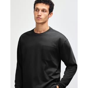A black super heavyweight t-shirt featuring a pocket, modelled by a person with curly hair. It has a logo displayed.