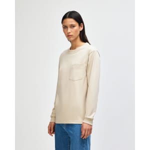 A long-sleeve beige t-shirt with a pocket and a crew neck, displayed by a model wearing olive green pants.
