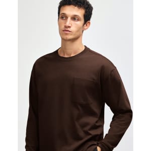 A model wears a brown American Apparel Super Heavyweight T-Shirt with a chest pocket. The image features a logo.