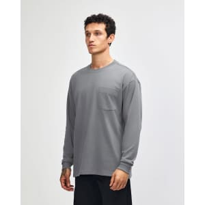 A man is wearing a grey long-sleeve t-shirt with a pocket, standing in a neutral backdrop. The shirt has a logo.