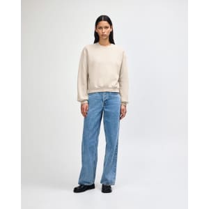 A women's crewneck sweatshirt in a light beige colour, styled with blue jeans and white shoes. It has a small logo.