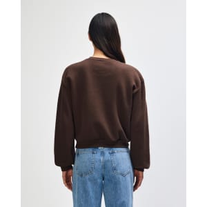 A women's crewneck sweatshirt in brown, featuring a relaxed fit and a logo. The model is shown from the back.