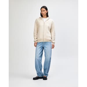 A unisex full zip hooded sweatshirt in beige, worn by a model standing in black cargo pants and sneakers.