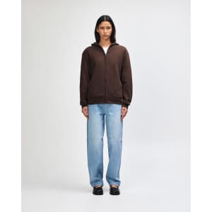 A unisex full zip hooded sweatshirt in brown, worn with light blue baggy jeans and sneakers. Features a logo.