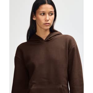A brown unisex hooded sweatshirt with a front pocket and a model adjusting the hood. It features a logo.