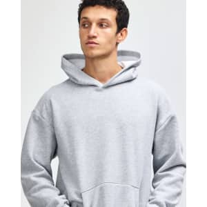 A grey unisex hooded sweatshirt with a front pocket and a logo. The model is standing with hands in pockets.