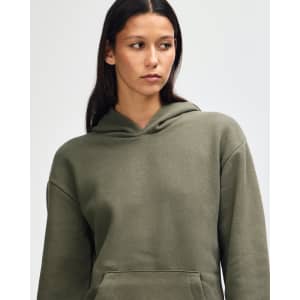 A unisex hooded sweatshirt in olive green with a front pocket and a logo. The model is wearing it casually.