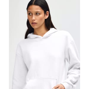 A unisex hooded sweatshirt in white, featuring a pocket and a logo on the front. The model adjusts the collar.