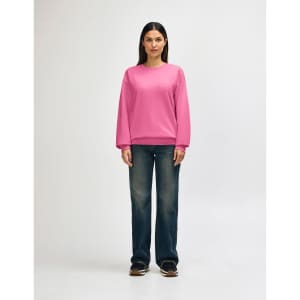 A woman in a pink Gildan Softstyle adult sweatshirt and blue jeans, adjusting her sleeve. The sweatshirt has a logo.