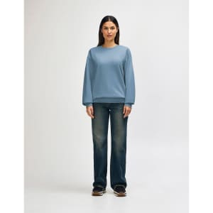 A woman stands wearing a light blue Gildan Softstyle sweatshirt paired with blue jeans and white sneakers.