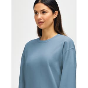 A blue Gildan Softstyle adult sweatshirt is worn by a model adjusting their sleeves, featuring a logo on the garment.