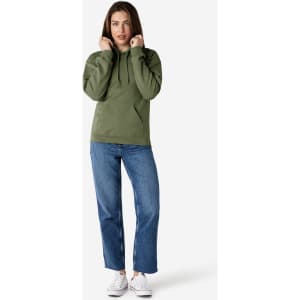 A woman stands wearing a green hooded sweatshirt and blue jeans, with her hands near the hood. The sweatshirt has a logo.