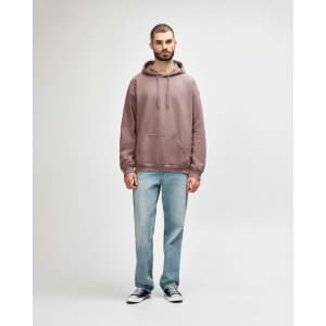 A woman wearing a dusty rose hooded sweatshirt paired with blue jeans and white shoes. The sweatshirt has a logo.