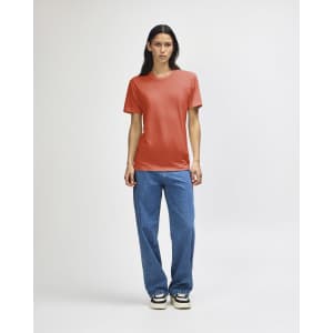 A woman stands wearing an orange American Apparel tri-blend T-shirt and blue denim jeans, featuring a logo.