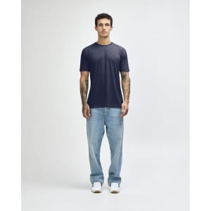 A model stands wearing a navy American Apparel Tri-Blend T-shirt and light blue jeans, with white shoes.