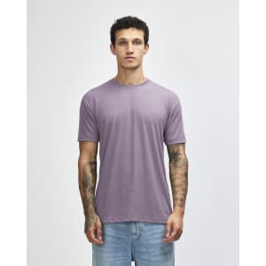 A man wearing a purple American Apparel tri-blend T-shirt paired with light blue jeans, featuring visible tattoos on his arms.