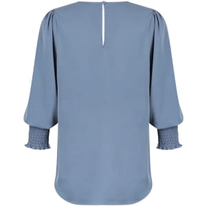 A blue blouse featuring long sleeves with smocked detail and a back keyhole opening. It has a branding logo.