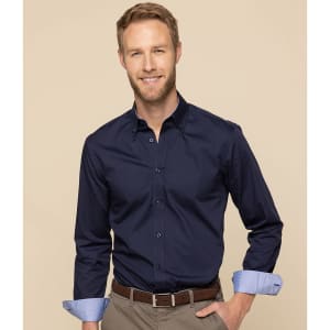 A man wearing a navy button-up shirt with rolled-up light blue cuffs, smiling against a neutral background.