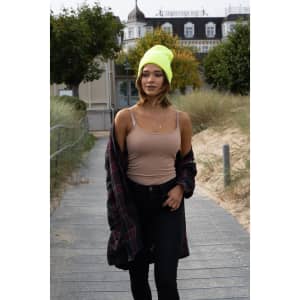A woman wearing a neon yellow beanie and a beige tank top walks along a wooden path surrounded by greenery.