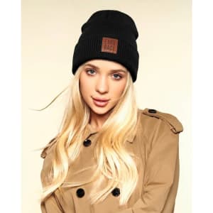 A black knitted beanie with a pom pom, featuring a branded patch, modelled by a person with long blonde hair.