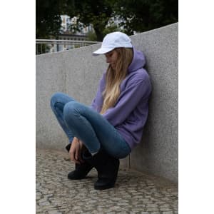 A woman wearing a white cap sits on a grey stone surface, dressed in a purple hoodie and blue jeans.