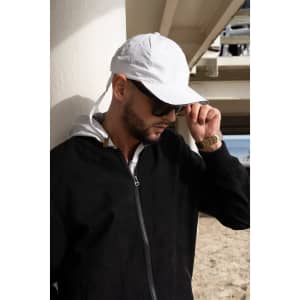 A white cap with a wind strap and clip, worn by a man in sunglasses and a black jacket, near a beach setting.