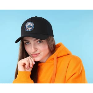 A woman wearing a black cotton twill cap with a buckle, paired with an orange hoodie. The cap features a logo.