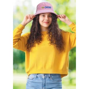 A pink terry towelling bucket hat with a logo, worn by a smiling person in a yellow sweatshirt against a green background.