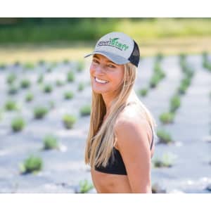A woman with long blonde hair wears a grey and black trucker cap, smiling outdoors in a field of plants.