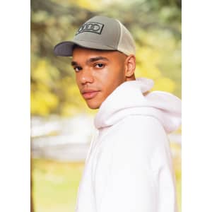 A man in a grey cap and white hoodie stands outdoors, showcasing a stylish look against a blurred natural background.