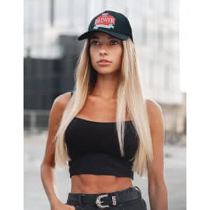 A woman with long blonde hair wearing a black tank top and cap with a logo stands outdoors.