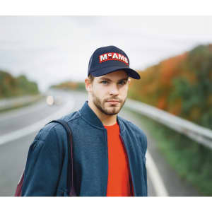 A man wearing a navy cap with a logo, a blue bomber jacket, and a red shirt, standing near a road with autumn trees.