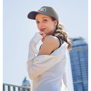 A woman wearing a black cap and white shirt poses outdoors, with a city skyline in the background.