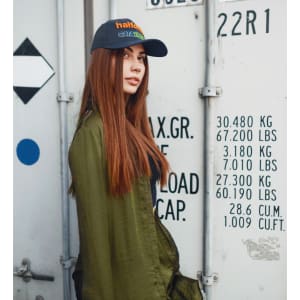 A woman with long brown hair wears a blue cap and a green jacket, standing against a shipping container background.