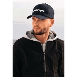 A navy cap with a logo, worn by a person in a grey hoodie and black jacket, set against a natural background.