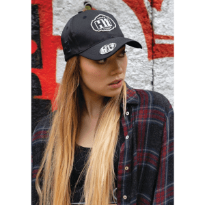 A black twill cap with a logo, worn by a model with long hair, styled with a plaid shirt in an urban setting.