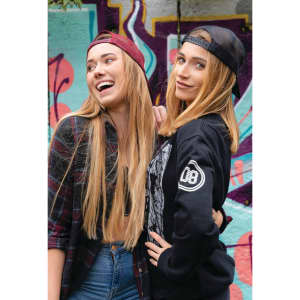 Two young women wearing caps, one in burgundy and the other in black, pose against a vibrant graffiti backdrop.