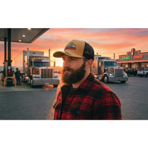A man with a beard wears a tan and black trucker cap, standing beside trucks at a gas station during sunset.