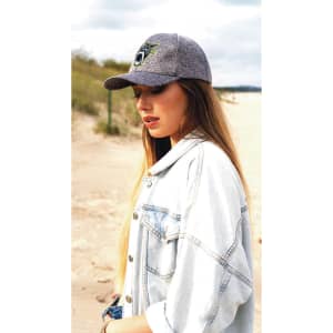 A woman wears a grey cap and a denim jacket, standing on a sandy beach with grass in the background.