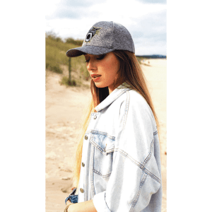 A grey cap worn by a woman with long hair, standing on a sandy beach, wearing a denim jacket.