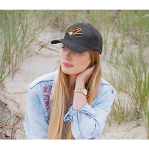 A woman with long hair wears a black cap and a denim jacket, sitting in a grassy area by the beach.