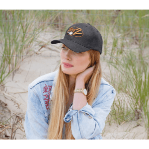 A black cap with a logo sits on a woman with long hair, wearing a denim jacket, surrounded by grass.